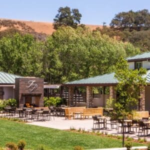 An outdoor patio with tables, chairs, and a fireplace at a building with a green roof and trees in the background.