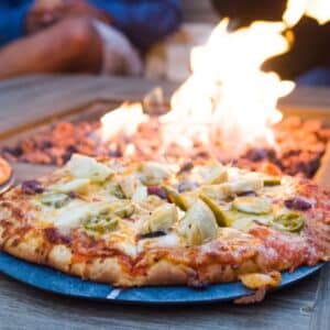 A pizza with artichoke hearts, jalapenos, and olives sitting near an outdoor fire pit.