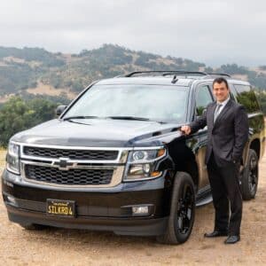 A man in a suit standing next to a black Chevrolet Suburban