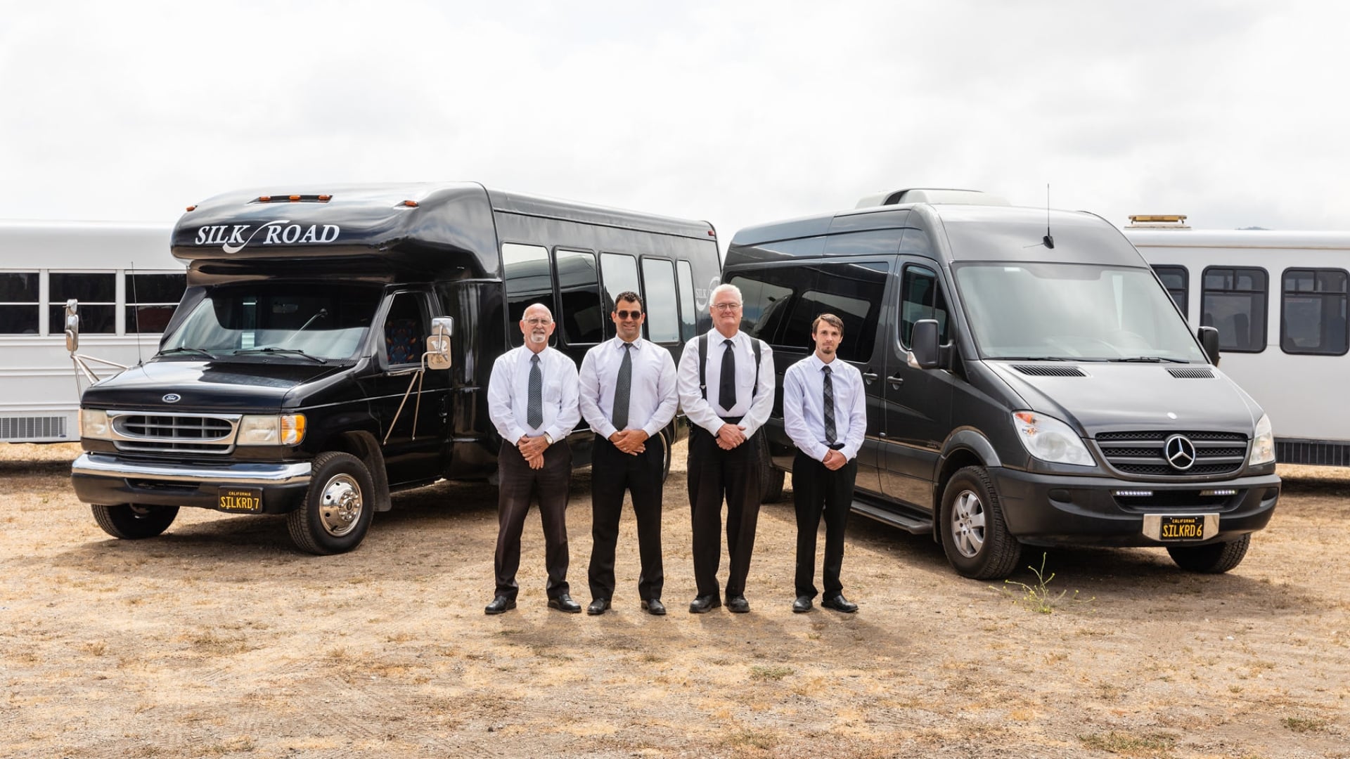 Four men in shirts and ties standing in front of a black Ford bus and a black Mercedes-Benz Sprinter bus